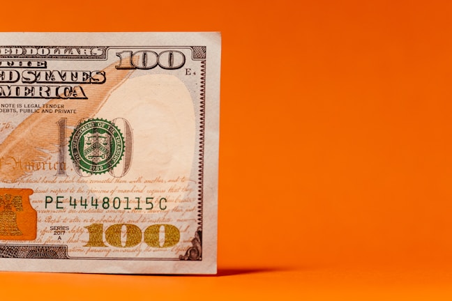 A close-up of a one hundred dollar bill partially placed against a vivid orange background, showing detailed features including the number 100 and parts of the official seal.