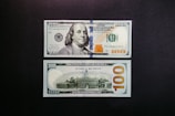 A US 100 dollar bill is displayed on a dark surface. Both the front and back of the bill are visible. The front features a portrait of Benjamin Franklin along with the denomination in numerals and text. The back shows an image of Independence Hall, with large numerals and a prominent '100' reading horizontally.