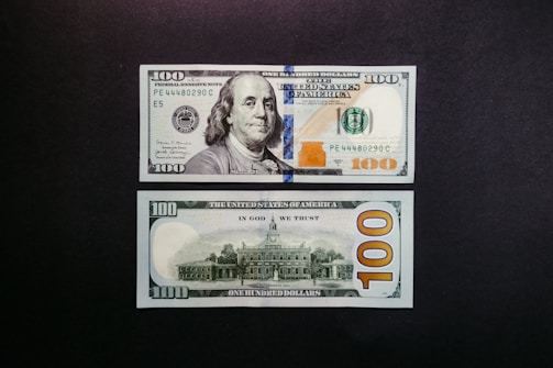 A US 100 dollar bill is displayed on a dark surface. Both the front and back of the bill are visible. The front features a portrait of Benjamin Franklin along with the denomination in numerals and text. The back shows an image of Independence Hall, with large numerals and a prominent '100' reading horizontally.
