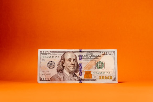 A US one hundred dollar bill positioned upright against a vibrant orange backdrop. The bill prominently features the portrait of Benjamin Franklin.