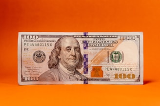 A US hundred-dollar bill is displayed against a vivid orange background. The bill features a portrait of Benjamin Franklin and includes typical security elements like a security strip.