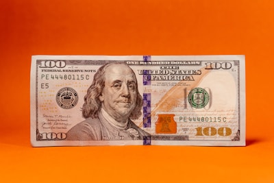 A US hundred-dollar bill is displayed against a vivid orange background. The bill features a portrait of Benjamin Franklin and includes typical security elements like a security strip.