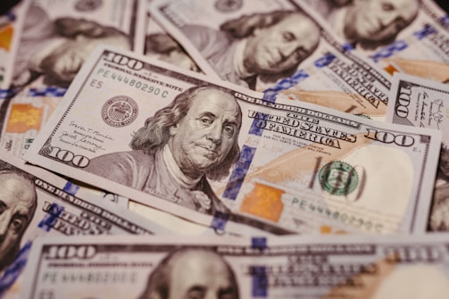A pile of U.S. one hundred dollar bills featuring the portrait of Benjamin Franklin, revealing intricate details of currency design including the Federal Reserve emblem and distinctive colors.