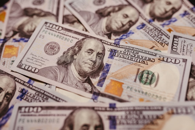 A pile of U.S. one hundred dollar bills featuring the portrait of Benjamin Franklin, revealing intricate details of currency design including the Federal Reserve emblem and distinctive colors.