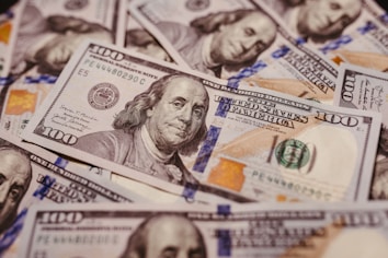 A pile of U.S. one hundred dollar bills featuring the portrait of Benjamin Franklin, revealing intricate details of currency design including the Federal Reserve emblem and distinctive colors.