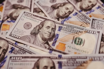 A pile of U.S. one hundred dollar bills featuring the portrait of Benjamin Franklin, revealing intricate details of currency design including the Federal Reserve emblem and distinctive colors.