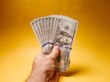 A hand holding multiple one hundred dollar bills against a plain yellow background. The bills are fanned out, showcasing Benjamin Franklin's portrait clearly.