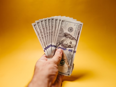 A hand holding multiple one hundred dollar bills against a plain yellow background. The bills are fanned out, showcasing Benjamin Franklin's portrait clearly.