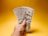A hand holding multiple one hundred dollar bills against a plain yellow background. The bills are fanned out, showcasing Benjamin Franklin's portrait clearly.