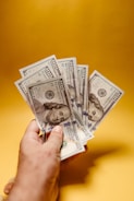 A hand is holding several one hundred dollar bills spread out against a bright, solid yellow background.