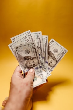 A hand is holding several one hundred dollar bills spread out against a bright, solid yellow background.