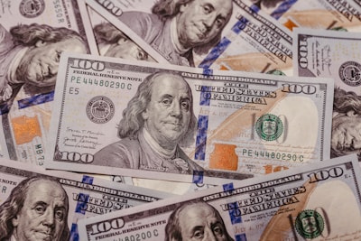 A pile of one hundred dollar bills featuring the portrait of Benjamin Franklin. The bills are arranged in an overlapping manner, showcasing both the front and portions of the back. Prominent security features, such as the blue security ribbon and color-shifting ink, are visible on the bills.
