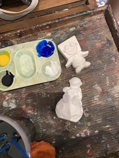 The image features a wooden table surface containing a small paint palette with black, yellow, blue, and white paints. Next to it is an unfinished, unpainted ceramic figure resembling a cartoonish character with large eyes and a wide smile. Another ceramic object below depicts a snowman sitting on a rounded base. The table is scattered with colorful paint spots from previous use.