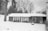 A snow-covered area featuring a house with a garage and porch. The garage door appears to be slightly damaged. Trees without leaves are visible in the background, and the entire scene is in black and white.