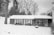 A snow-covered area featuring a house with a garage and porch. The garage door appears to be slightly damaged. Trees without leaves are visible in the background, and the entire scene is in black and white.