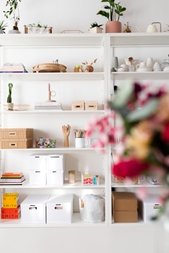 A neatly organized white shelving unit filled with various decorative items and storage boxes. The shelves contain green plants, woven baskets, books, art supplies including colored pencils, and stacked storage boxes. The background is minimal and bright, with a soft focus on vibrant flowers in the foreground.