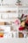 A neatly organized white shelving unit filled with various decorative items and storage boxes. The shelves contain green plants, woven baskets, books, art supplies including colored pencils, and stacked storage boxes. The background is minimal and bright, with a soft focus on vibrant flowers in the foreground.