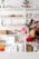 A neatly organized white shelving unit filled with various decorative items and storage boxes. The shelves contain green plants, woven baskets, books, art supplies including colored pencils, and stacked storage boxes. The background is minimal and bright, with a soft focus on vibrant flowers in the foreground.