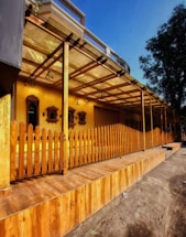 A wooden deck area is enclosed by a vertical wooden fence. The structure has a roof supported by wooden beams and a covered walkway. The background features yellow walls with decorative wooden frames and various hanging lamps. There is also a tree to the right side providing some greenery.