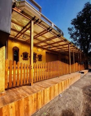 A wooden deck area is enclosed by a vertical wooden fence. The structure has a roof supported by wooden beams and a covered walkway. The background features yellow walls with decorative wooden frames and various hanging lamps. There is also a tree to the right side providing some greenery.