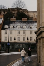 Two people dressed in winter clothing walk along a snow-dusted street towards a building labeled Waterside Hotel. The street is partially covered with snow, and the building has a quaint, traditional look with small windows and a sloped roof. Bare trees are visible in the background.