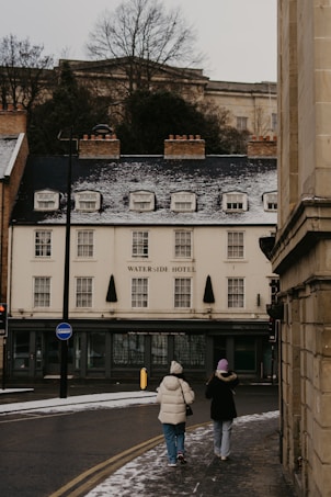 Two people dressed in winter clothing walk along a snow-dusted street towards a building labeled Waterside Hotel. The street is partially covered with snow, and the building has a quaint, traditional look with small windows and a sloped roof. Bare trees are visible in the background.