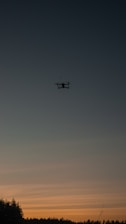 A drone hovers against a backdrop of a darkening sky during sunset. The sky is transitioning from deep blue at the top to warm orange hues near the horizon. Silhouettes of trees and vegetation are visible at the bottom of the image, adding depth and natural contrast.