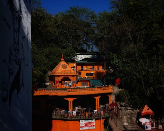 A vibrant orange building stands against a backdrop of lush green trees. The structure features intricate designs and a pointed dome on top. People can be seen on the balconies, and there are decorative elements and signs in a language containing Sanskrit characters. The sky is clear and blue, providing a vivid contrast to the orange and green.