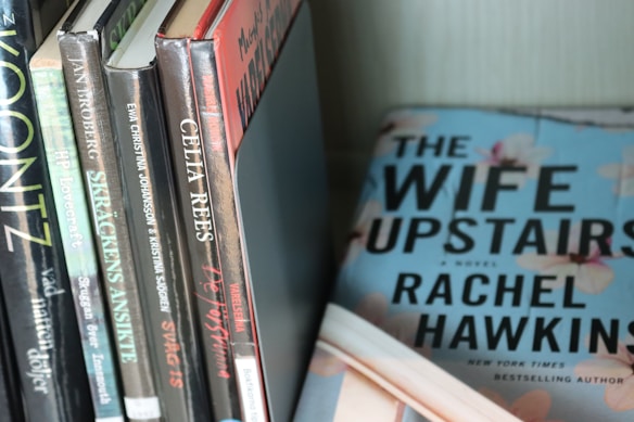 A collection of books is neatly arranged on a shelf. The titles include works by authors such as Lovecraft, Koontz, and Celia Rees. A hardcover book titled 'The Wife Upstairs' by Rachel Hawkins is prominently displayed, lying flat with a blue cover featuring large bold lettering and floral designs.