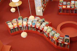 A public library section with red shelves filled with colorful children's books arranged alphabetically in an orderly manner. The flooring is red carpeted and overhead spherical pendant lights cast a soft glow. The ambiance is warm and inviting, perfect for reading or browsing books.