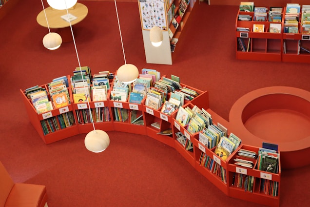 A public library section with red shelves filled with colorful children's books arranged alphabetically in an orderly manner. The flooring is red carpeted and overhead spherical pendant lights cast a soft glow. The ambiance is warm and inviting, perfect for reading or browsing books.
