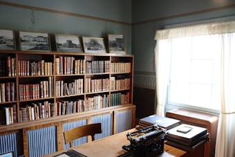 A cozy study room featuring a large bookcase filled with assorted books. On top of the shelves, framed black and white photographs are displayed. A wooden desk in the foreground holds an old-fashioned typewriter and a stack of books. Sunlight streams in through a large window with light curtains, creating a warm and serene atmosphere.