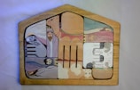 A wooden jigsaw puzzle with various painted sections. The sections depict abstract and somewhat childlike drawings of biblical figures and animals, including a person with a halo, another with a crown, and some animals that resemble sheep. The pieces fit into a house-shaped frame, each part forming a segment of the overall picture.