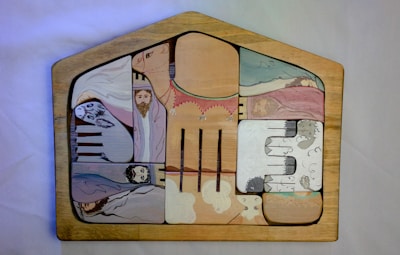 A wooden jigsaw puzzle with various painted sections. The sections depict abstract and somewhat childlike drawings of biblical figures and animals, including a person with a halo, another with a crown, and some animals that resemble sheep. The pieces fit into a house-shaped frame, each part forming a segment of the overall picture.