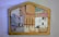 A wooden jigsaw puzzle with various painted sections. The sections depict abstract and somewhat childlike drawings of biblical figures and animals, including a person with a halo, another with a crown, and some animals that resemble sheep. The pieces fit into a house-shaped frame, each part forming a segment of the overall picture.