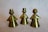 Three shiny brass figurines are displayed, each resembling a stylized, abstract human form. They have round heads and conical bodies, with one wearing a crown, indicating a royal theme. The surface of each figure reflects light with a yellowish tint.