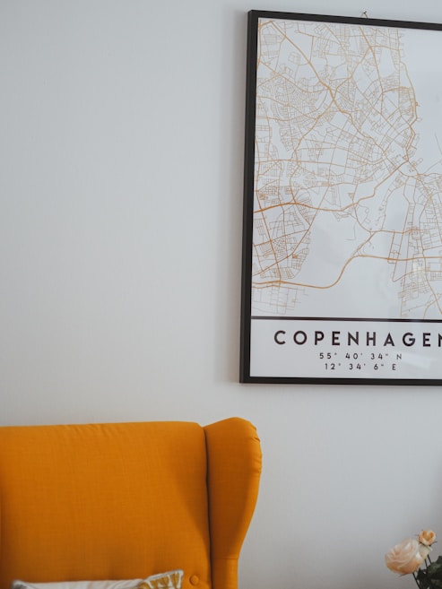 A cozy interior with a mustard yellow armchair partially visible next to a map of Copenhagen framed on a light-colored wall. The cushion and a pale flower bouquet add subtle detail to the scene.
