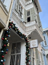 A charming, historic building adorned with festive decorations and lights. Traditional architecture with wooden shutters and intricate balcony railings reflects a vintage style. The facade is decorated with garlands and colorful ornaments, creating a cheerful, festive atmosphere. A sign with the words 'Rita Moda' hangs in front.