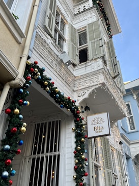 A charming, historic building adorned with festive decorations and lights. Traditional architecture with wooden shutters and intricate balcony railings reflects a vintage style. The facade is decorated with garlands and colorful ornaments, creating a cheerful, festive atmosphere. A sign with the words 'Rita Moda' hangs in front.