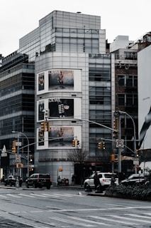 An urban streetscape featuring a modern multi-story building with large glass windows. Prominent advertising billboards are displayed on the building, showing fashion-related content. Several vehicles are on the road, and a few pedestrians are visible on the sidewalks. Traffic lights and street signs are part of the busy city scene.