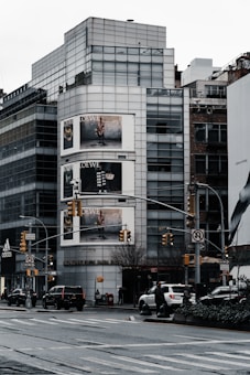An urban streetscape featuring a modern multi-story building with large glass windows. Prominent advertising billboards are displayed on the building, showing fashion-related content. Several vehicles are on the road, and a few pedestrians are visible on the sidewalks. Traffic lights and street signs are part of the busy city scene.