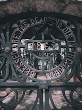 An ornate iron gate with intricate designs stands as the focal point. The words 'Blessed are the dead who die in the Lord' are engraved along the circular arch of the gate. A black ribbon is tied to the center, just above the year '1862,' adding a solemn touch. The background reveals a cobblestone pathway leading into a cemetery surrounded by tall, dense trees.