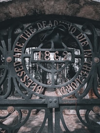 An ornate iron gate with intricate designs stands as the focal point. The words 'Blessed are the dead who die in the Lord' are engraved along the circular arch of the gate. A black ribbon is tied to the center, just above the year '1862,' adding a solemn touch. The background reveals a cobblestone pathway leading into a cemetery surrounded by tall, dense trees.