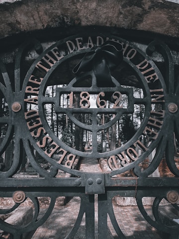 An ornate iron gate with intricate designs stands as the focal point. The words 'Blessed are the dead who die in the Lord' are engraved along the circular arch of the gate. A black ribbon is tied to the center, just above the year '1862,' adding a solemn touch. The background reveals a cobblestone pathway leading into a cemetery surrounded by tall, dense trees.
