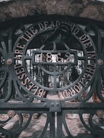 An ornate iron gate with intricate designs stands as the focal point. The words 'Blessed are the dead who die in the Lord' are engraved along the circular arch of the gate. A black ribbon is tied to the center, just above the year '1862,' adding a solemn touch. The background reveals a cobblestone pathway leading into a cemetery surrounded by tall, dense trees.