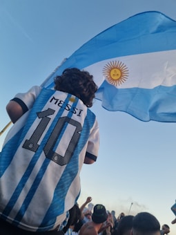 A person wearing a number 10 jersey with 'Messi' on the back stands in a crowd, holding a large Argentine flag against a clear blue sky.