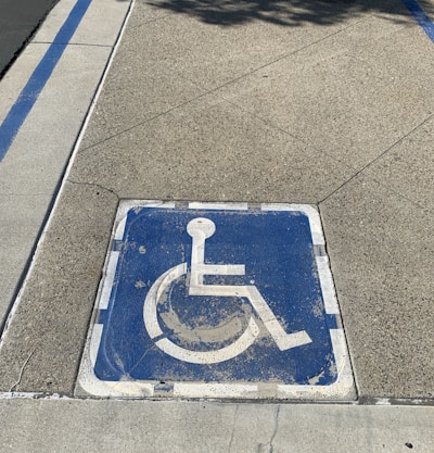 A weathered blue wheelchair symbol painted on a square area of pavement, indicating a designated parking space for individuals with disabilities. It is bordered by faded white lines, and the surrounding surface is composed of textured concrete.