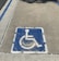 A weathered blue wheelchair symbol painted on a square area of pavement, indicating a designated parking space for individuals with disabilities. It is bordered by faded white lines, and the surrounding surface is composed of textured concrete.