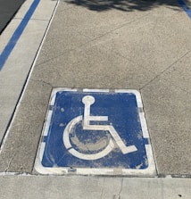 A weathered blue wheelchair symbol painted on a square area of pavement, indicating a designated parking space for individuals with disabilities. It is bordered by faded white lines, and the surrounding surface is composed of textured concrete.