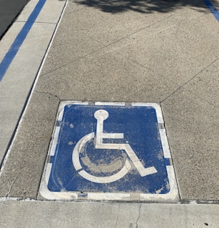 A weathered blue wheelchair symbol painted on a square area of pavement, indicating a designated parking space for individuals with disabilities. It is bordered by faded white lines, and the surrounding surface is composed of textured concrete.